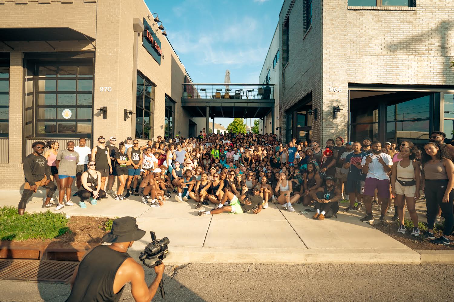 A large group of runners posing for a group photo in front of a brick building on a warm sunny day