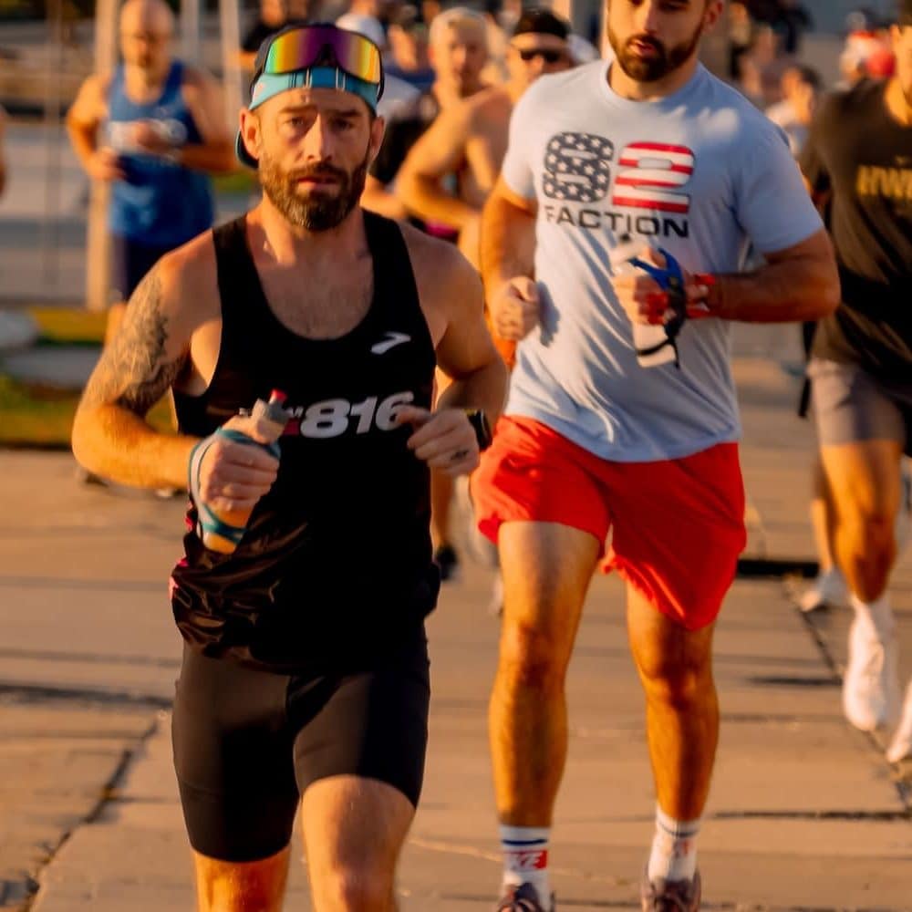 A male runner in a black tank top and shorts leading a race with other runners behind him, running on pavement