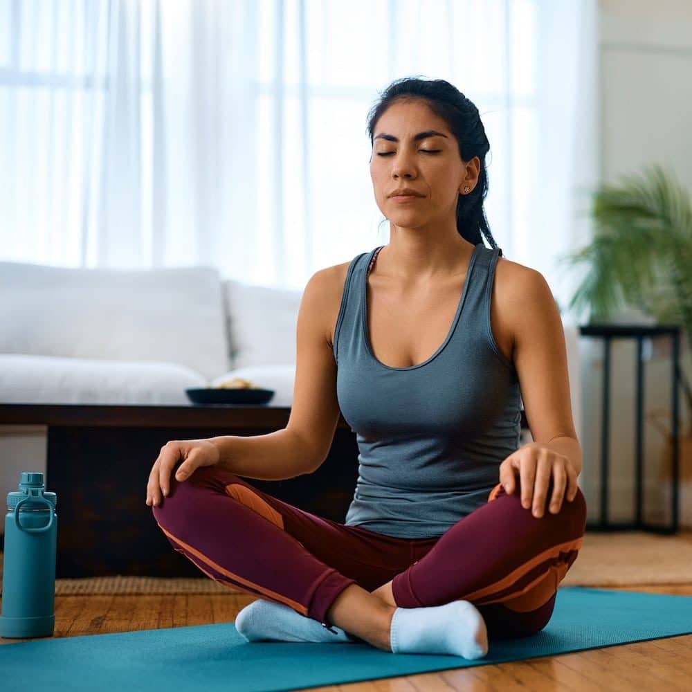 Female athlete in lotus pose practicing yoga with eyes closed in her living room with furniture in the background